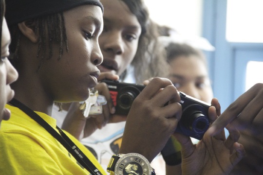 Two high school girls holding cameras on which they are learning how to edit a video.