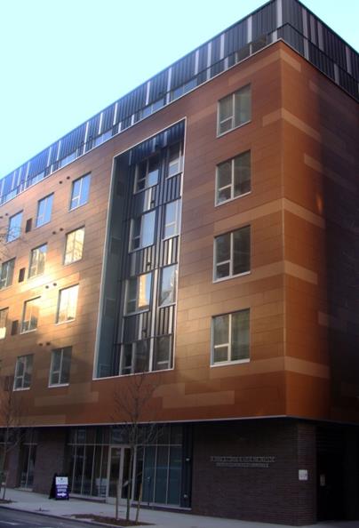 Photograph taken at street level from across the street, showing the front façade of the building. The first floor exterior is finished with marble and includes a series of glass doors. Floors two through four, covered in wood paneling, protrude from the building except for an indented accent feature above the building entrance. The top floor of the flat-roofed building is set back from the lower floors and covered in corrugated metal.