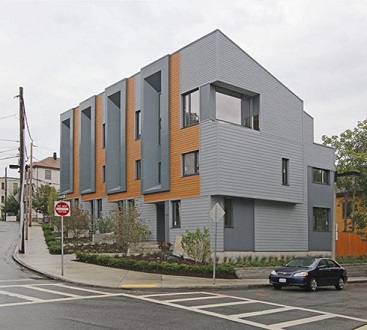 Photograph taken at street level from across the intersection of the front and side facades of a three-story townhouse building. A small front yard landscaped with ground cover, plants, and shrubs sits between the sidewalk and the front doors of the four units.