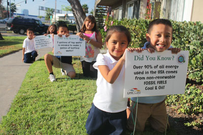 Three pairs of children on the grounds of City Gardens Apartments holding yard signs with facts about energy and water use.