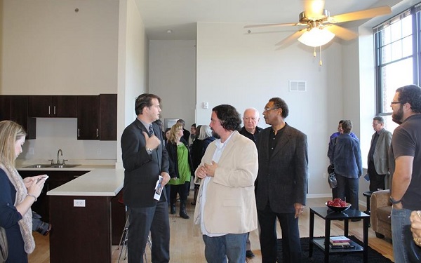 Photograph of several people touring one of the residential units and looking over the kitchen area and living area with large windows.