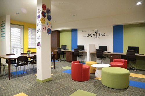 Photograph of the development’s community room, which is furnished with a work table and lounge chairs in the middle of the room and a row of computer work stations along a wall.