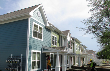 Photograph taken at street level of the front façade of approximately 10 two-story townhouse residences. Two workers are preparing the front yards for landscaping.