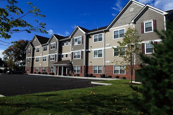 Photograph of a façade of a three-story residential building, with brick on the first floor and siding above. A one-story portico marks the building’s entrance, and six gables mark the roofline. In the foreground are a parking lot with grass and trees on the perimeter. Small bushes line the parking lot in front of the building.