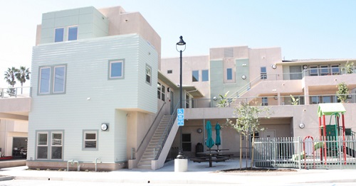 Photograph taken at street level of a three-story apartment building also showing part of a playground and an outdoor sitting area.