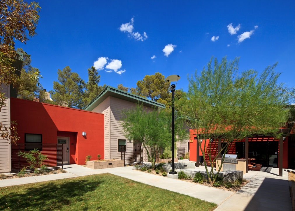 Ground level photograph of the courtyard landscaped with grass, shrubs, and trees. A pergola covers a portion of the courtyard; nearby are a grill, picnic tables, and a low semi-circular wall for sitting. Buildings line two sides of the courtyard in the background; entrances to two residential units are in the photograph along one of the walls.