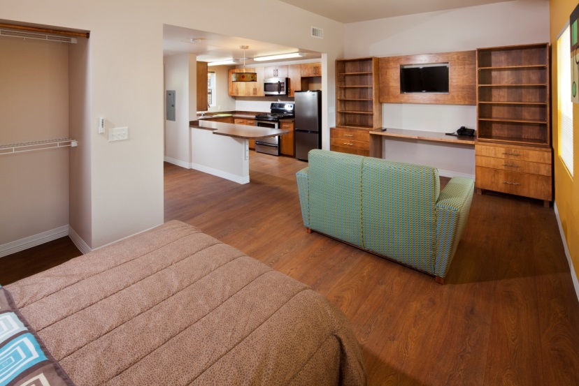 Ground-level photograph of the interior of a studio unit taken from the sleeping area, with a bed in the foreground, looking through the living area with a couch and a built-in storage/desk unit and toward a fully equipped kitchen.