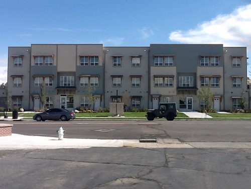 Photograph of the front façade of a flat-roofed, three-story building made of a variety of materials. The building is set back from the street by a wide lawn. Cars are parked along the street in the foreground.