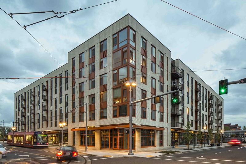 Photograph taken from street level at an intersection, showing two street fronts of Gray’s Landing, a six-story building with commercial uses on the first floor and residences above.