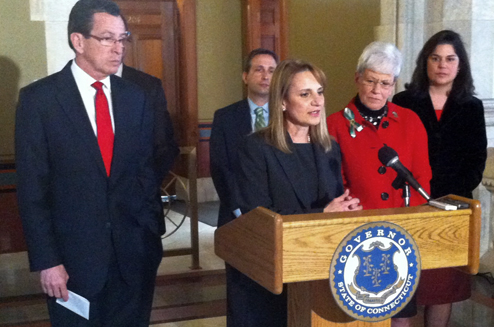 Photograph of six people in a Connecticut government office. Standing at a lectern is Evonne Klein, newly appointed commissioner of housing for the state. Governor Dannel P. Malloy and Lieutenant Governor Nancy Wyman stand on either side of her.