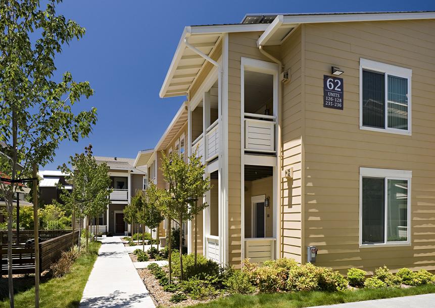 Exterior image of low-rise apartment buildings with balconies and patios overlooking a sidewalk and surrounding vegetation. 