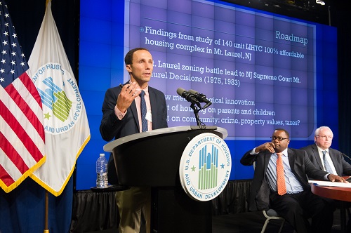 A panelist speaks from a podium bearing the HUD logo on stage. The American Flag and a HUD flag are visible to the left of the speaker, and a presentation screen and the speaker’s table can be seen in the background. Two seated panelists, turned towards the speaker, are visible on the right side of the image.