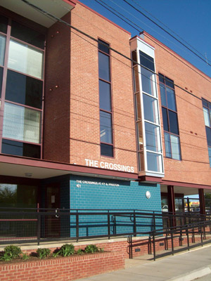 Photograph taken at street level of a portion of the front façade of a four-story building showing the entrance to the building and part of a switchback ramp providing access to the entrance approximately three feet above the sidewalk. A sign reading “The Crossings” in cut-out letters sits above the first floor near the building entrance.