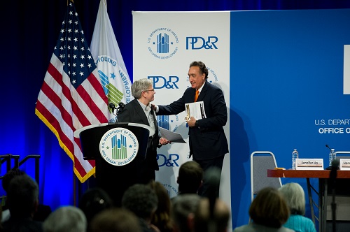 At the center of the image are Henry Cisneros, who is walking up to the podium, and Jennifer Ho, who is turning away from the podium. In the background are the American Flag, HUD Flag, and a PD&R backdrop.