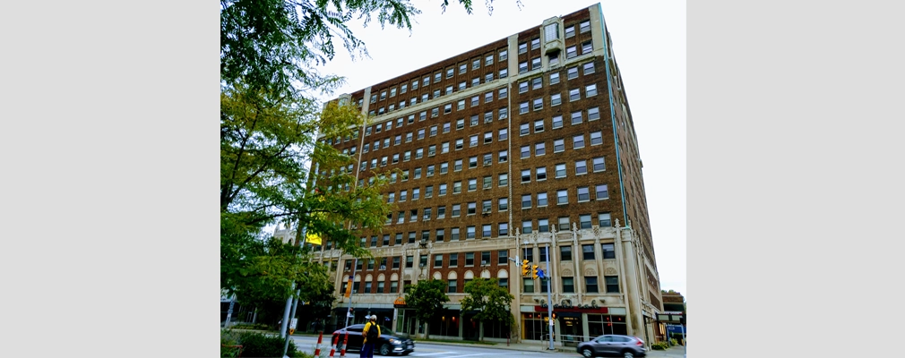 A 13-story, brick-and-concrete building on a city street corner, with street trees in front of the building.