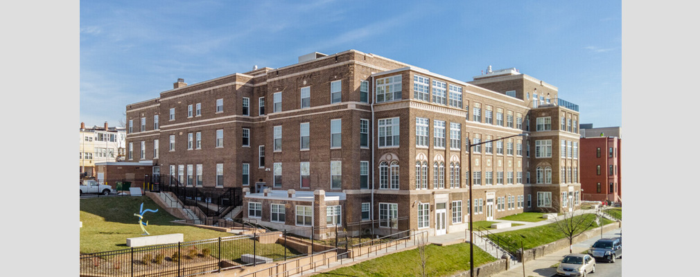 Corner view of a four-story brick building with a pocket park on one side and a public sidewalk in the foreground.