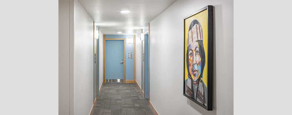 Photograph of an interior hallway with a tribal symbol next to an apartment number and a painting of a Native American woman in the foreground.