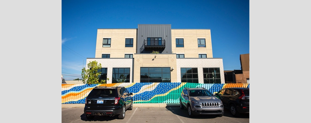 Photograph of a parking lot with parked cars in front of a mural and a three-story apartment building in the background.