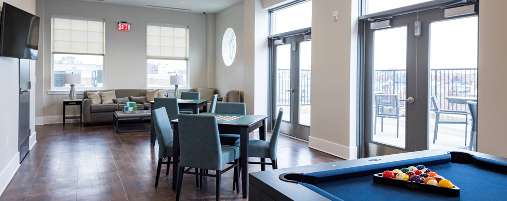 Interior of a community activity room with a pool table in the foreground, various seating arrangements and a wall-mounted television. 