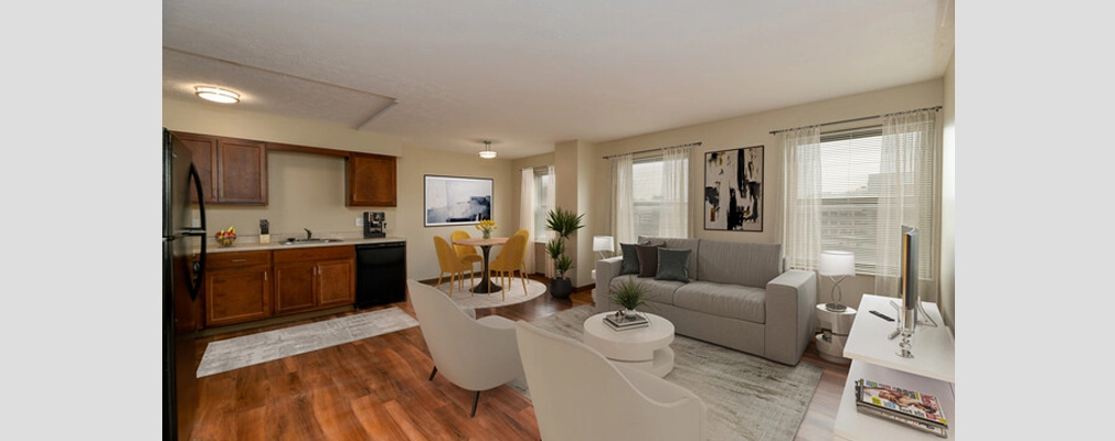  Interior of an open-floor plan apartment with living area seating and windows along one wall and kitchen in the background.