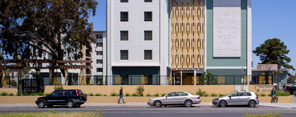 Street view of a six-story building behind a fence and concrete wall with a road with cars in the foreground.