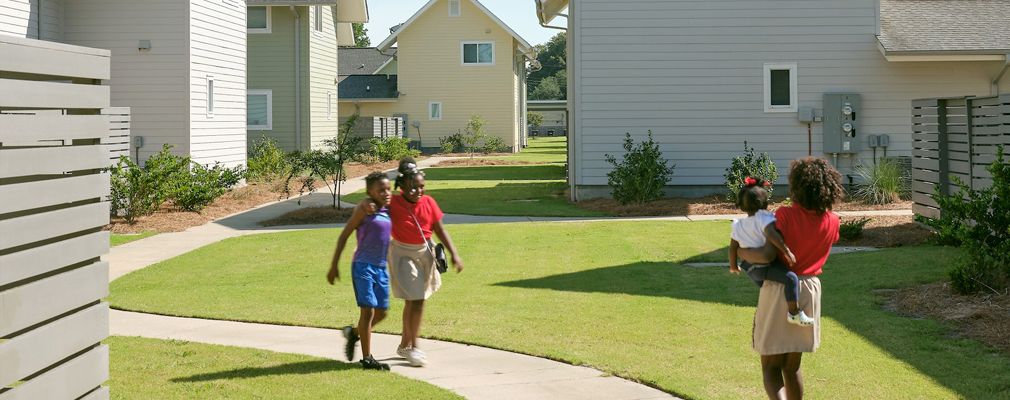 Three kids on a walking path with grass and two-story buildings on either side.