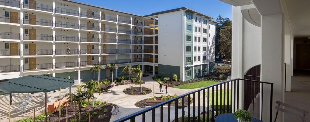 A balcony-level view of a landscaped courtyard framed by a six-story building, with a balcony railing, table, and two chairs in the foreground.