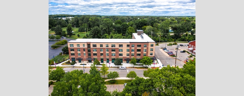 Four-story residential building surrounded by parking lots and large trees.