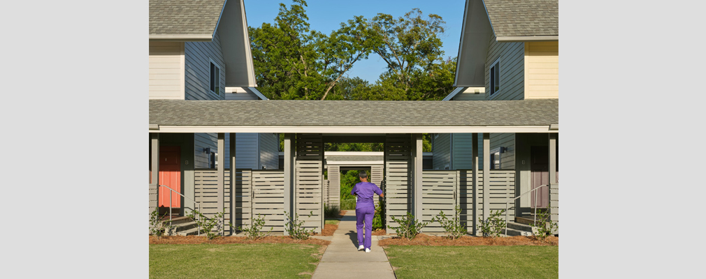 A person walking through an opening in a wall connecting two buildings with lawn in the foreground. 