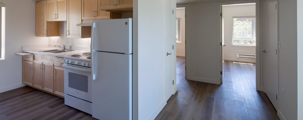 Photograph of a residential unit with a kitchen in the foreground and a hallway leading to two unfurnished rooms in the background.