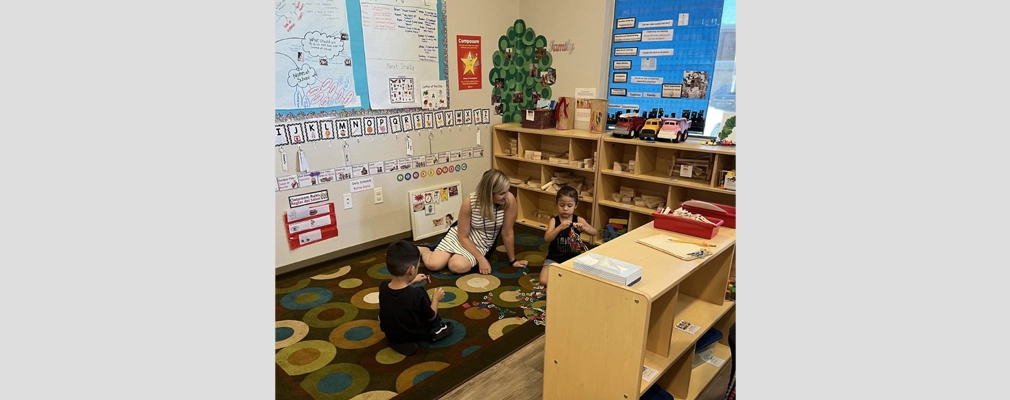 An adult and two small children sitting on a classroom floor, playing with toys. 