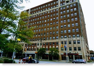 A 13-story, brick-and-concrete building on a city street corner, with street trees in front of the building.