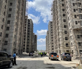 Construction site with tall, unfinished buildings on both sides of a road, under a partly cloudy sky. Several cars and people are visible on the road.