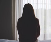 Backside of a woman looking out a window while sitting on a bed.