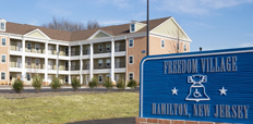 A three-story brick apartment building sits on a grassy lawn. A sign reads "Freedom Village: Hamilton, New Jersey."