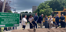 Federal, State, and local-Syracuse stakeholders at Interstate 81 carrying shovels dig into a pile of dirt. A large sign on the left reads "PROJECT FUNDED BY THE Bipartisan Infrastructure Law - BUILDING A BETTER AMERICA."
