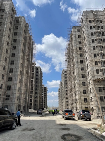 Construction site with tall, unfinished buildings on both sides of a road, under a partly cloudy sky. Several cars and people are visible on the road.