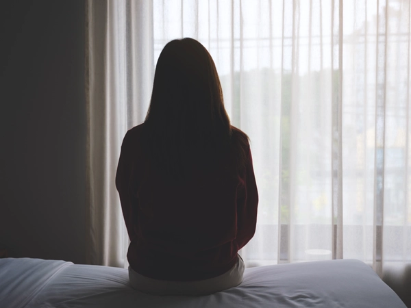 Backside of a woman looking out a window while sitting on a bed.