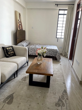 A room featuring a bed, sofa, and wooden coffee table with flowers. A window with curtains allows natural light in.