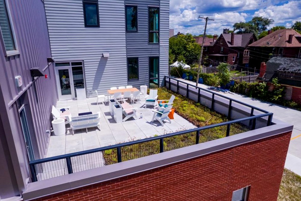 A landscaped rooftop terrace containing chairs, couches, and tables with a street and detached houses in the background.