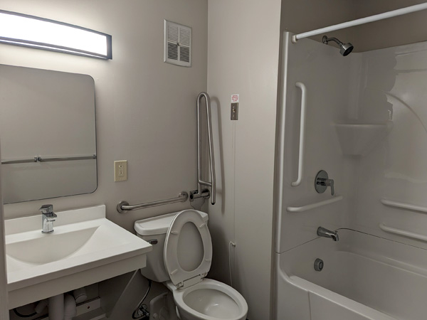 A new, clean bathroom with a tub and shower, toilet, sink, mirror, and several grab bars. 