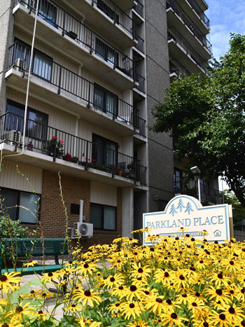 Exterior of a tall brick-and-concrete apartment building with a sign that reads, 'Parkland Place' and a patch of yellow flowers.
