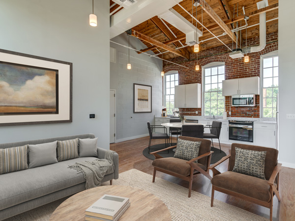 A spacious apartment interior with large windows, a modern kitchen, dining and seating areas, and exposed wood ceiling beams.