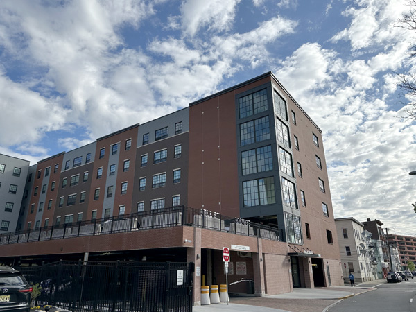 A modern red brick apartment building stands on a residential street. An older-looking three-story building is visible next to it.