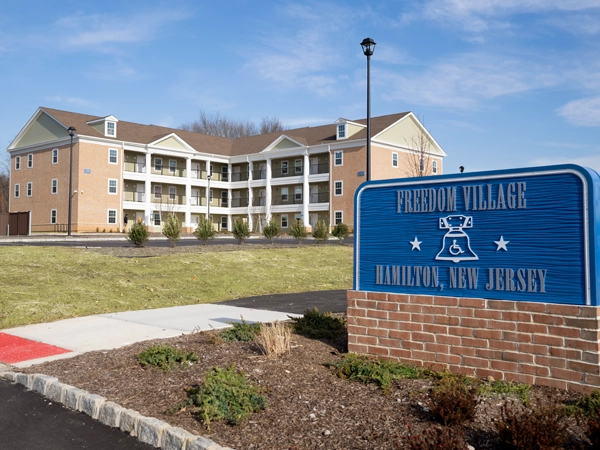 A three-story brick apartment building sits on a grassy lawn. A sign reads "Freedom Village: Hamilton, New Jersey."