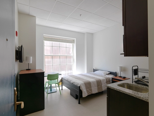 The interior of a studio apartment with a window, desk, chair, bed, nightstand, dresser, and kitchenette. 