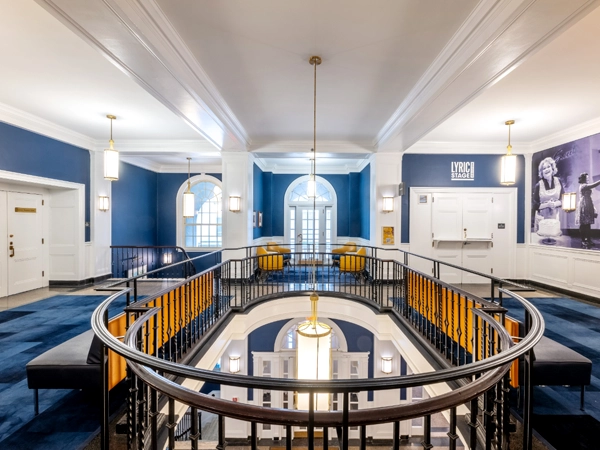 Floor lounge with a central oblong railing lined with benches and an opening to the floor below in the center; in the background is a seating area with chairs, two sets of double doors, and an inscription at the top of the back right wall that reads, "LYRIC STAGE BOSTON."