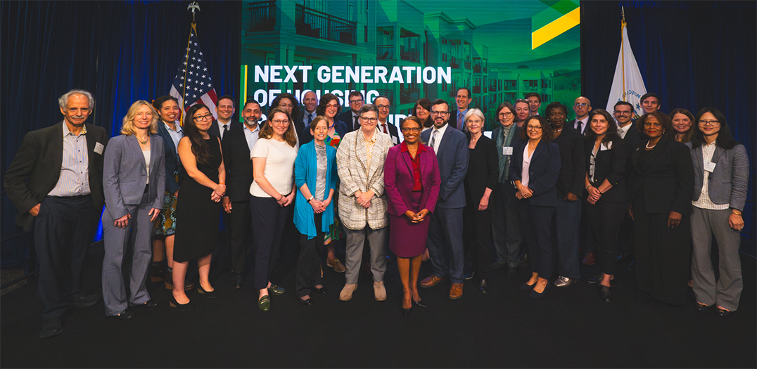 Group photo of PD&R staff and housing scholars standing in front of a large screen that reads "Next Generation of Housing Policy Roundtable."