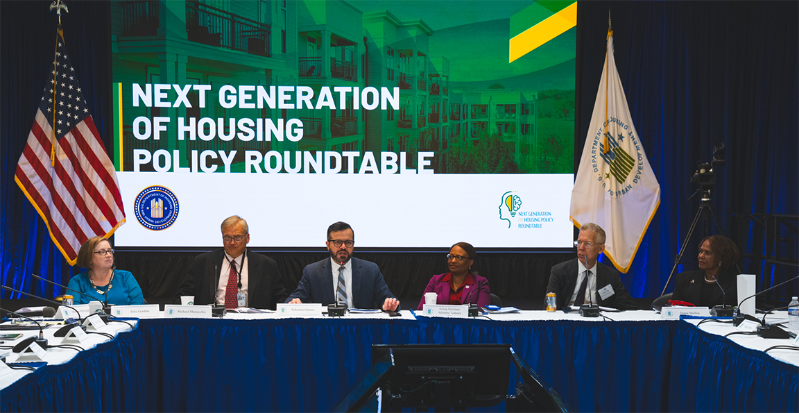 Participants sit around a table during the roundtable discussion. A large screen behind them reads "Next Generation of Housing Policy Roundtable."