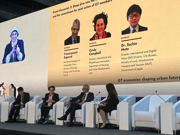 During a panel discussion, speakers sit in front of a large screen showing speaker names and titles.